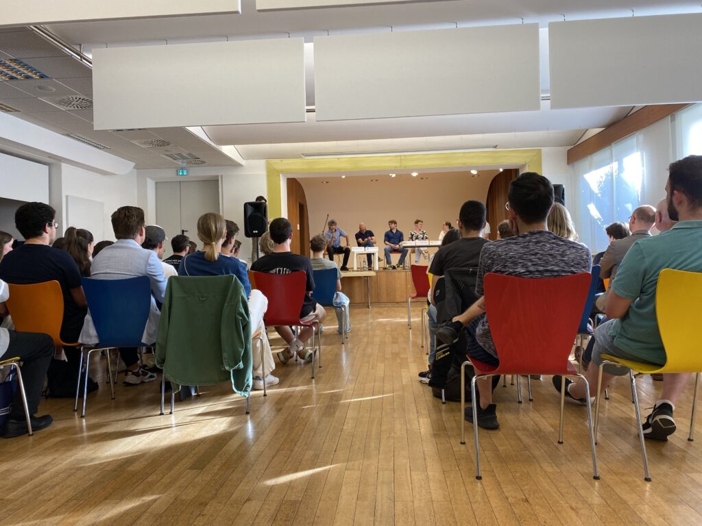 Podiusmdiskussion in Aula der Pestalozzi-Schule mit Personen auf der Bühne und Publikum sitzend davor.